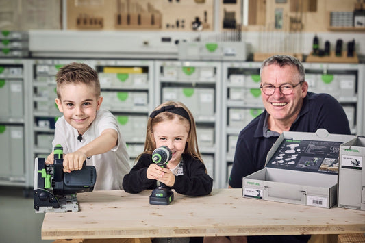 Festool TY-TSC Spielzeug Akku-Tauchsäge mit Vater und zwei Kindern, Akku und Zubehör auf Holztisch vor Systainer-Wand.